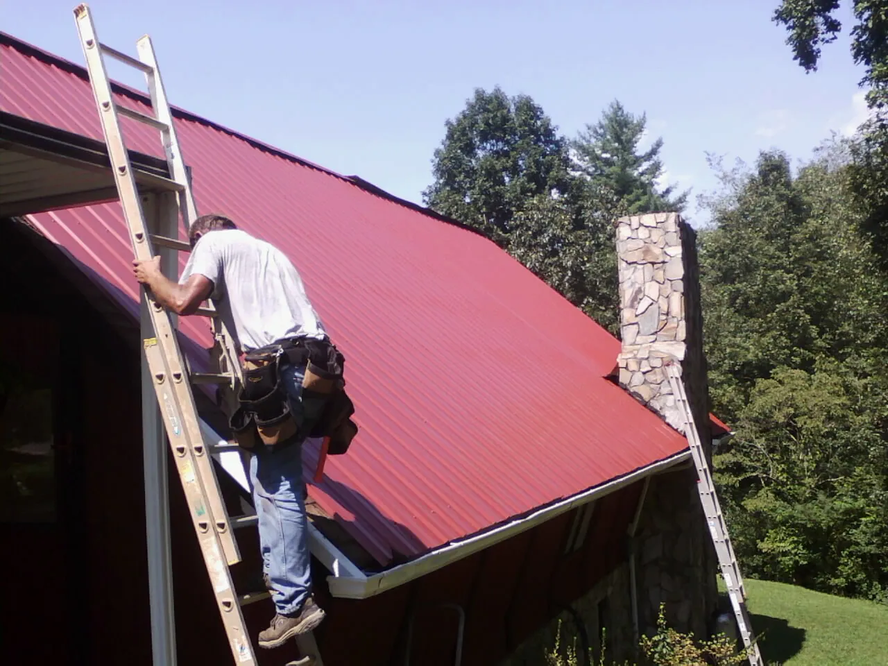 Roofer on a ladder beside a red metal roof and stone chimney — Soffit & Fascia Repair by Roofing Brothers in Mullan